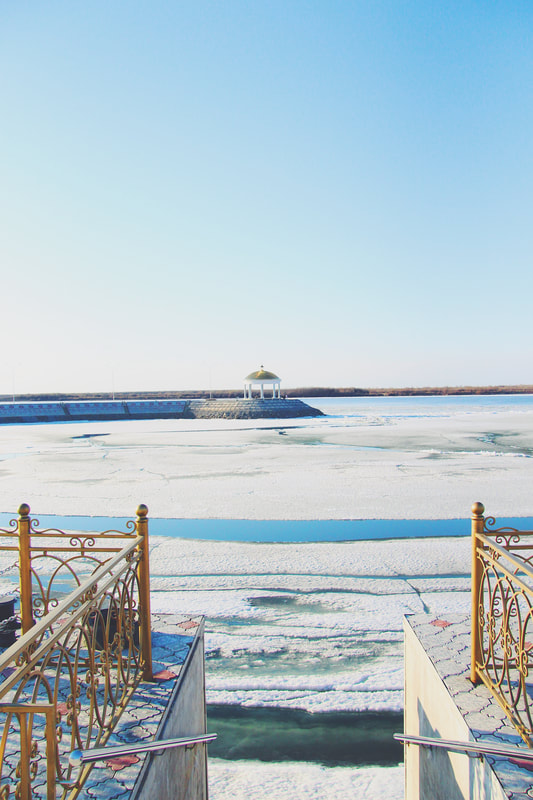 A gold railing is seen beside a staircase pointed at a gazebo which is visible in the distance. Ice is slowly melting on top of the water surrounding the gazebo. This photo was taken in Russia.