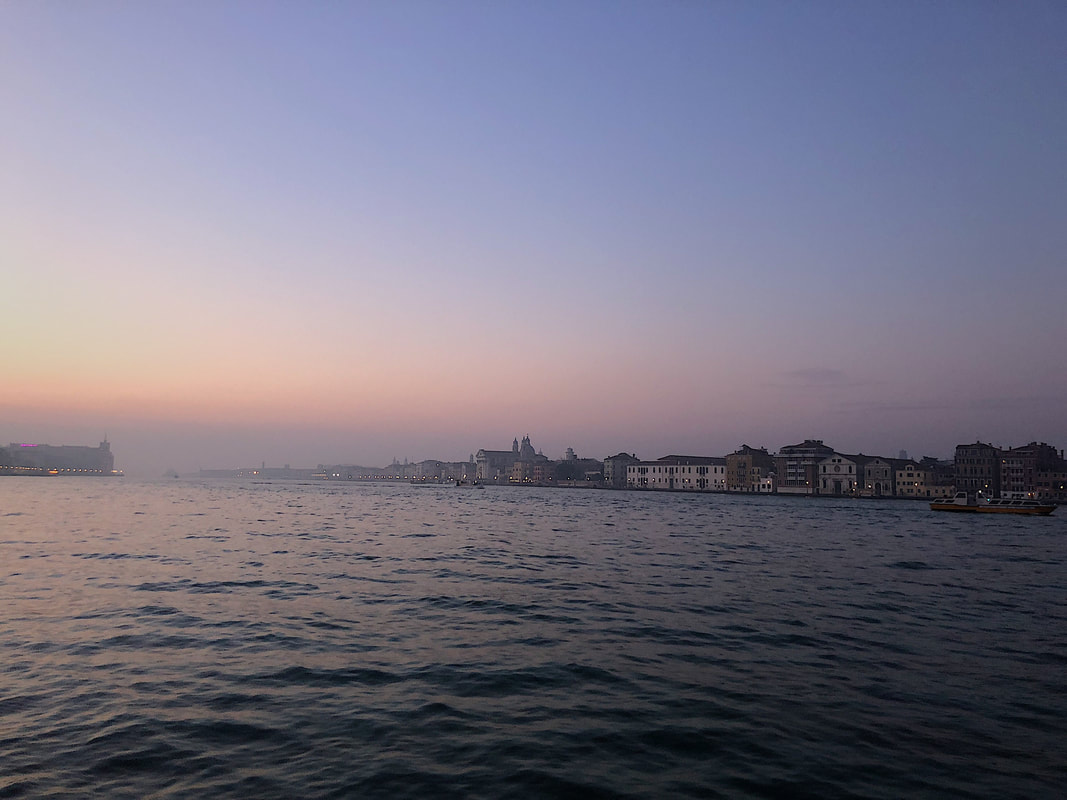 A purple sky is shown over Venice, Italy. Water occupies most of the image but buildings are visible in the distance. 