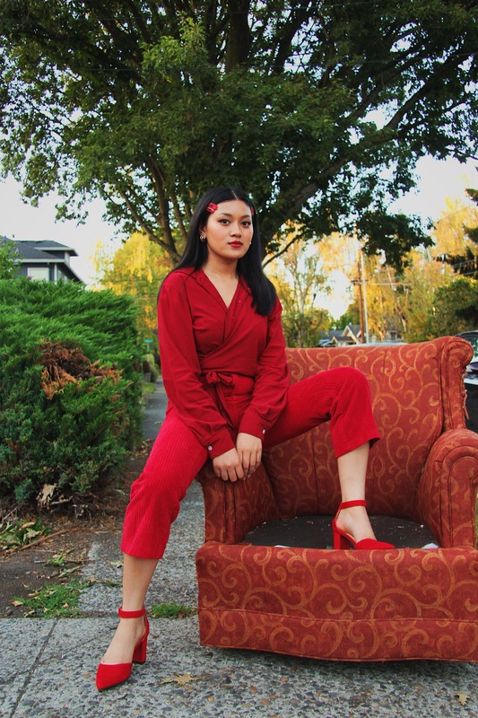 A woman dressed in all red sits with one leg up and one leg down on a chair. The chair is in the middle of a sidewalk of a suburban neighborhood.