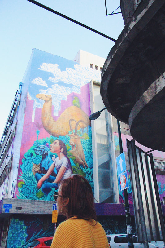 A woman in a yellow top (wearing sunglasses) is seen walking the streets of Tel Aviv. The camera points up in the direction of a mural on a building that depicts a women sitting while looking up, surrounded by animals such as a camel.