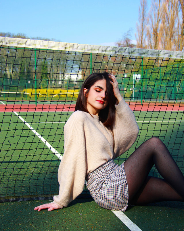 A woman sits on a tennis court beside the net. She touches her hair while leaning back on her other arm. She is wearing a sweater, skirt, and tights.