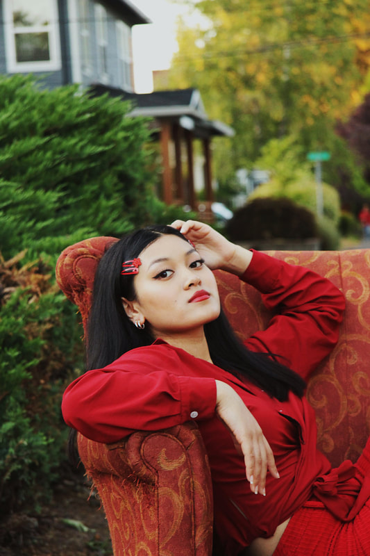 A woman in all red clothing reclines in a large chair that's outside in front of a bush and home.