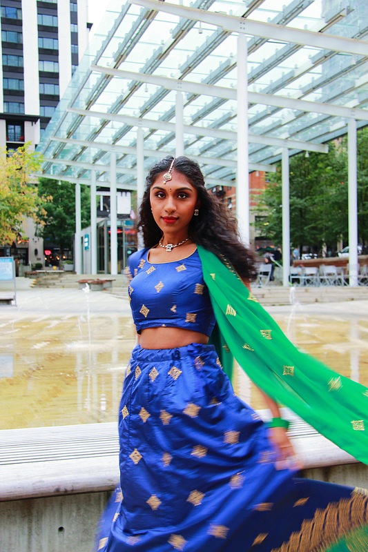 A woman twirls in her blue and green sari. She stands in front of a fountain.