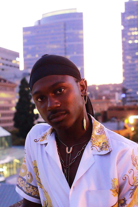 A man stares directly at the camera in front of downtown Portland. The buildings look purple. He is wearing a silk top with a few necklaces, earrings, and a durag.