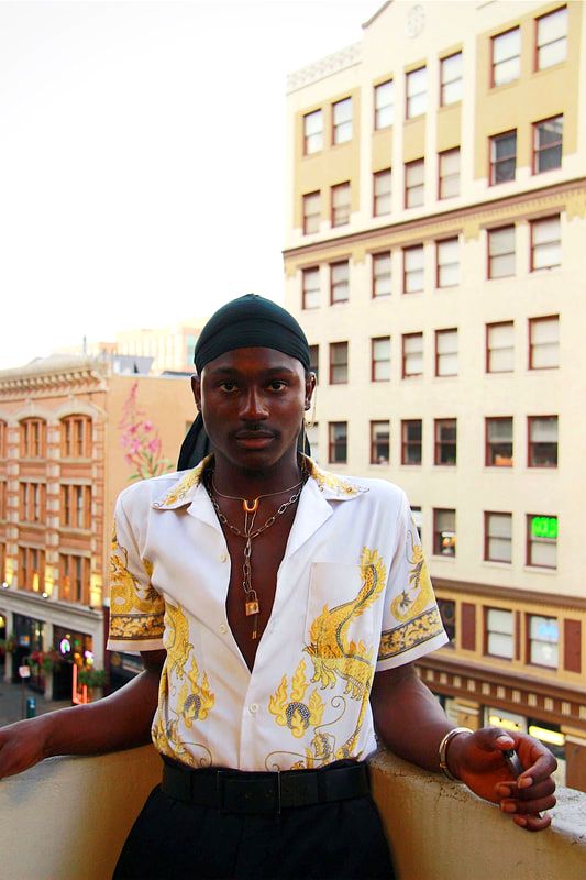 A man leans back on a wall which looks down on a main street in Portland. The photo includes many yellow tones. He is wearing black bottoms, a silk top, a durag, and jewelry.