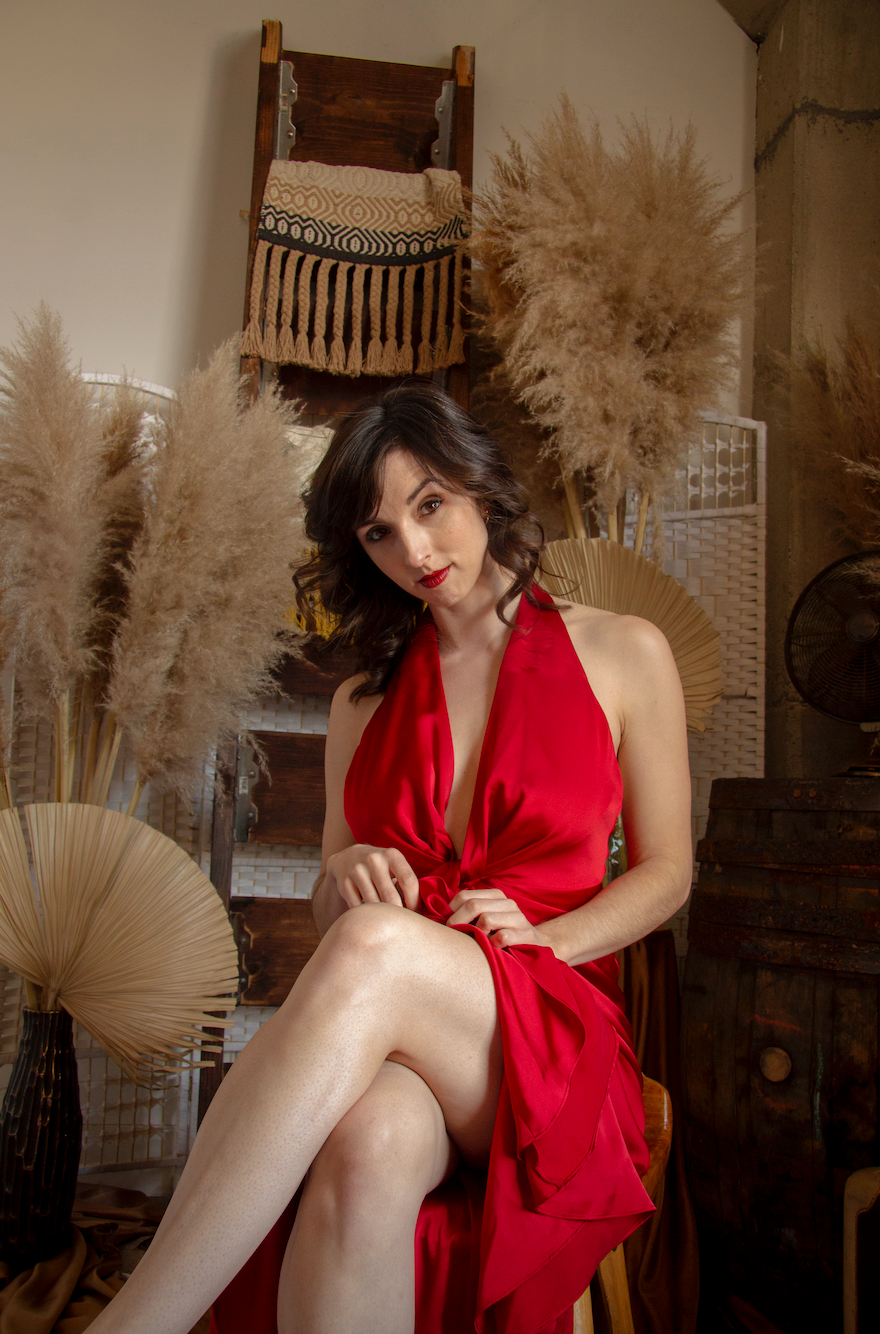 A woman in a long red dress is seen cross legged while sitting on a stool. Behind her is various props including dried out plants and a ladder with a cloth hanging on it.