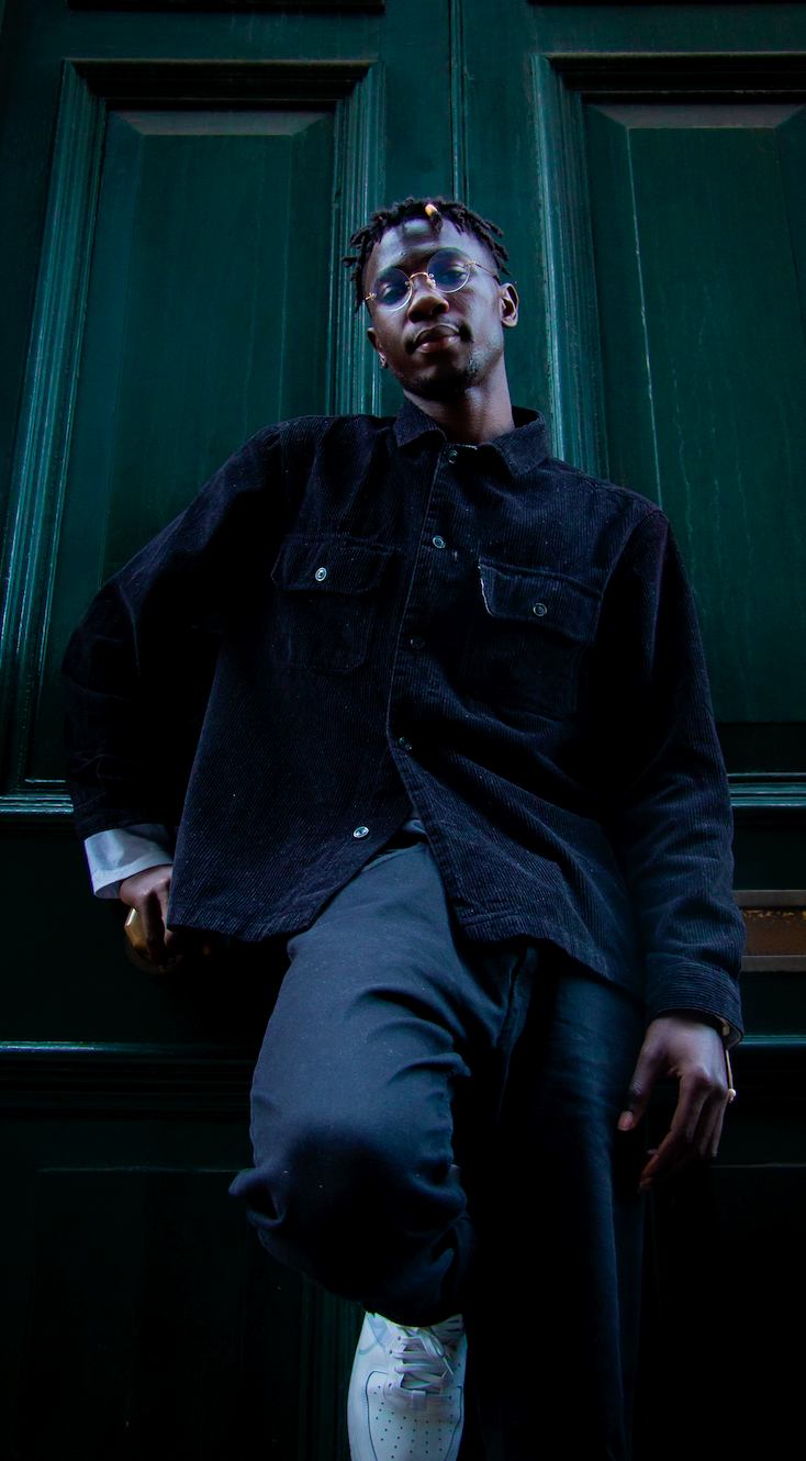 A man looks down at the camera while leaning on a wall. The tones of the wall and his clothing are dark.