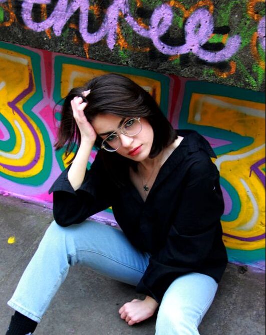 A woman that appears to be stressed leans on her hand while staring up at the camera and sitting down on the ground. Behind her is a bright, graffitied wall.