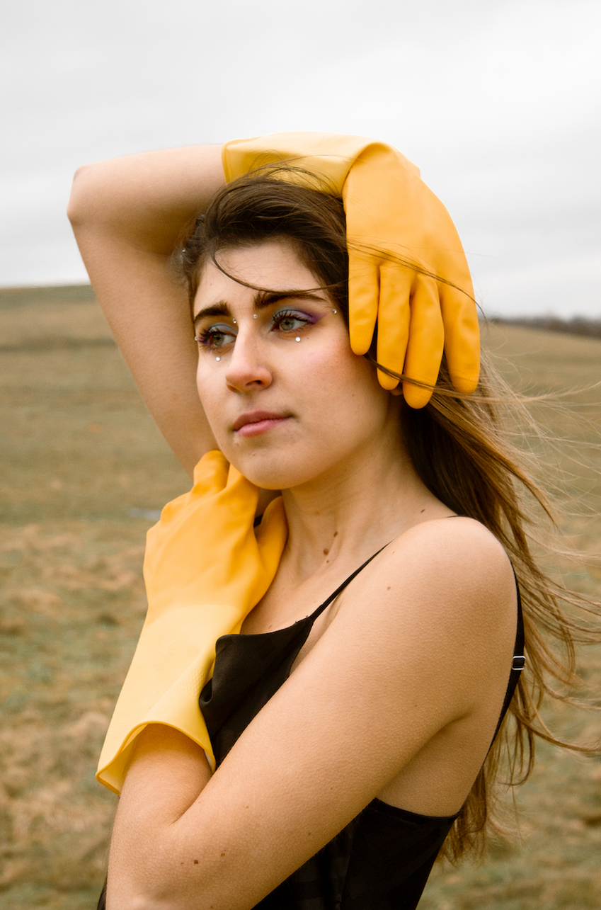 A woman stairs off into the distance while standing in a field in a black dress and yellow dishwasher gloves.  Her makeup includes sparkles and vibrant eyeshadow. The sky is gray. 