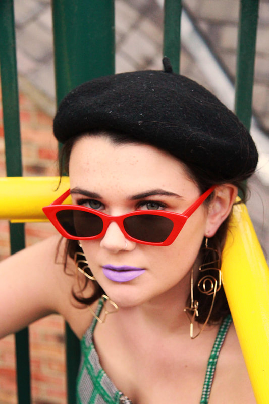 Image of a woman staring directly at the camera. She is wearing bright red glasses, a plaid top, earrings in the shape of a face, and a beret while leaning on a yellow railing.