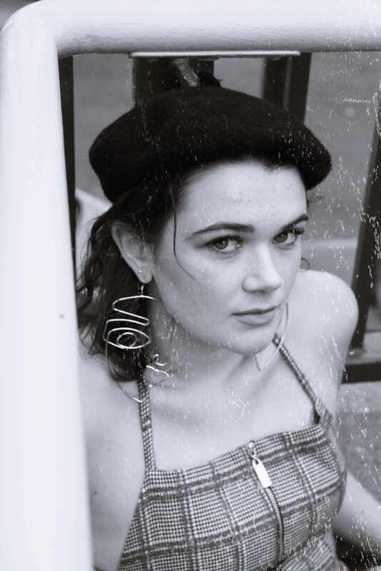 A woman in a black and white photo looks up at the camera while leaning on railing. She is wearing wire earrings, a beret, and a checkered top that zips up.