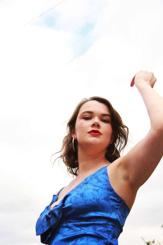 A woman in a blue dress and red lip looks down at the camera. One hand of hers is in the sky.