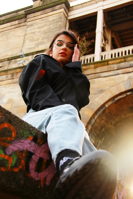 A woman touches her glass while sitting on top of a graffitied wall. A bright light shines behind her. The camera is angled from below her shoe.