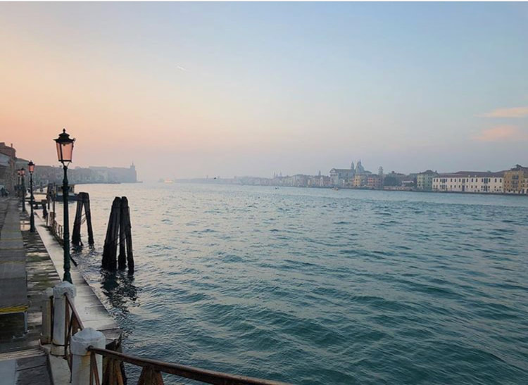 Image of the water surrounding Venice. The sky is a pastel color. A lamp post is visible to the left. Buildings are shown across the water.