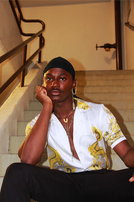 A man is shown staring off into the distance. This is a flash photo and his shadow is reflected behind him. He wears a durag, a silk top, and various jewelry including necklaces and a swooping earring.