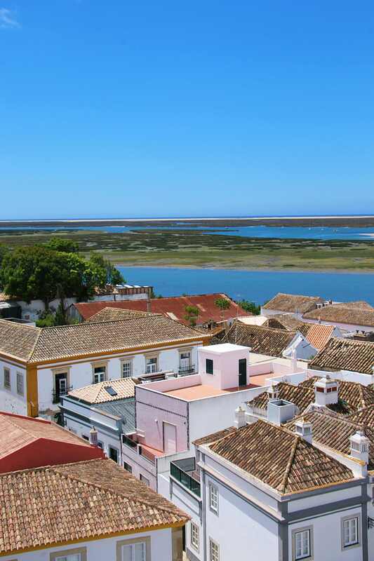 The rooftops of Faro, Portugal are shown. In the distance is a body of water and green trees to the left.