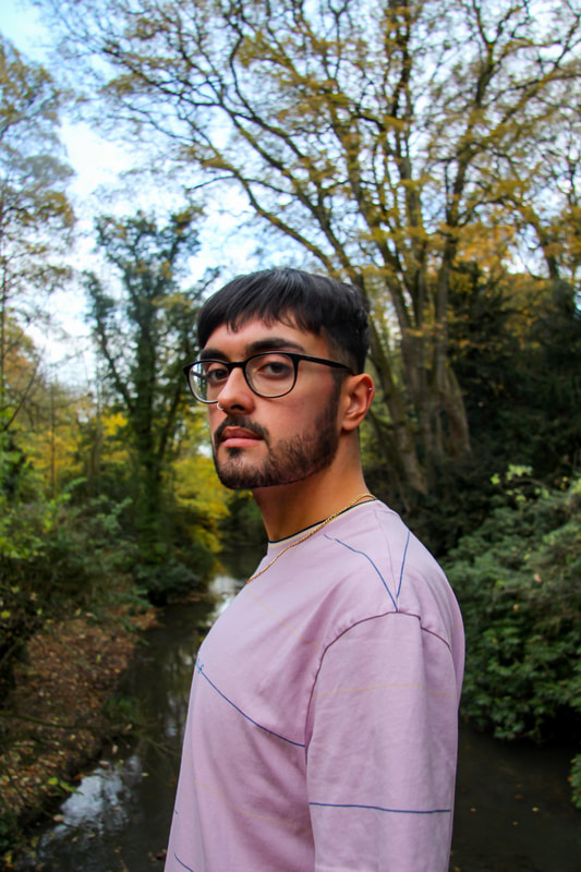 A man stares intensely into the camera in front of a forested background. He is wearing glasses and a purple top.