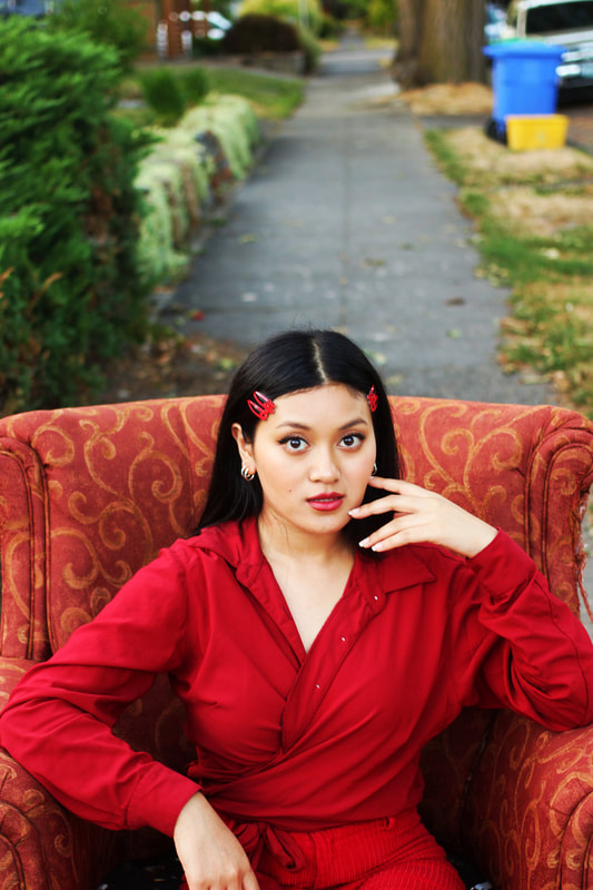Image of a woman sitting on a large chair in the middle of a sidewalk. Four clips pin her hair back and she's wearing an all red outfit.