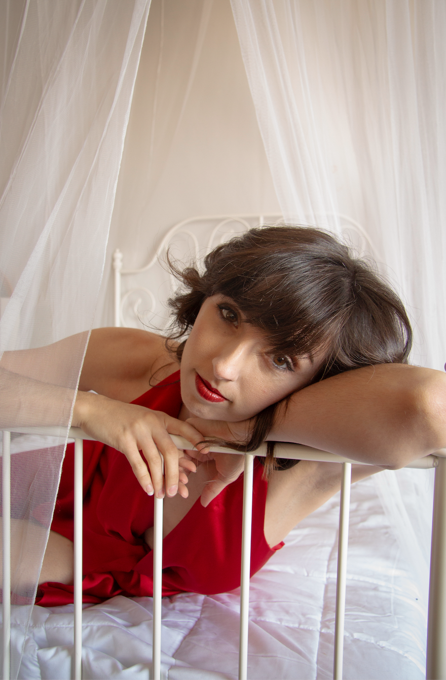 A woman leans on a bed railing while looking softly at the camera. She is wearing a red lip and dress. Above the bed hangs white curtains. 