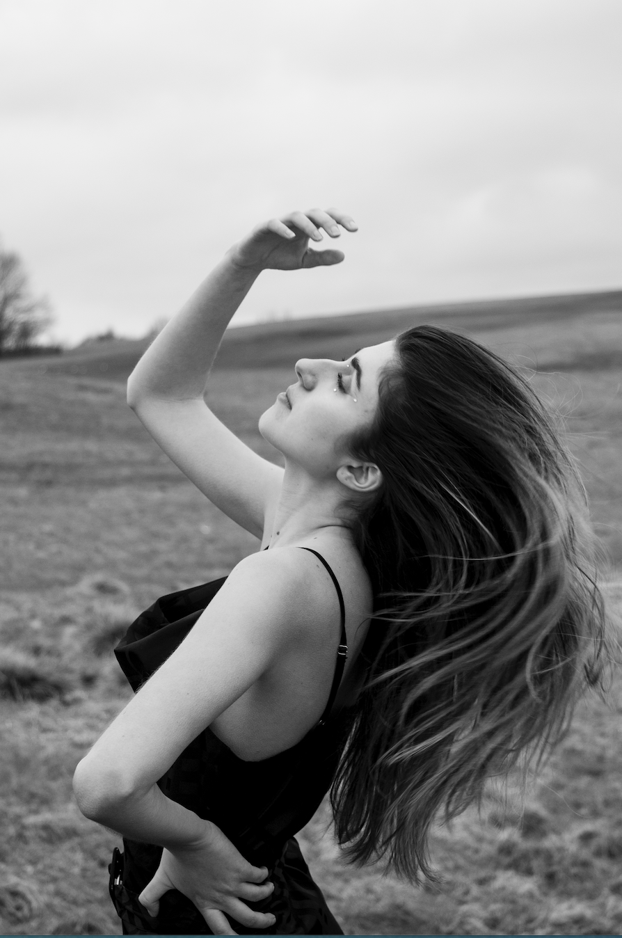 A woman in a black and white photo flips her hair back. She is standing in a field and her eyes are closed. She wears a long dress and her makeup includes sparkles around her eyes.